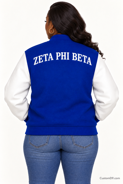 Black woman modeling Zeta Phi Beta royal blue body and white sleeve premium varsity jacket, back view; arched organization name across upper back.